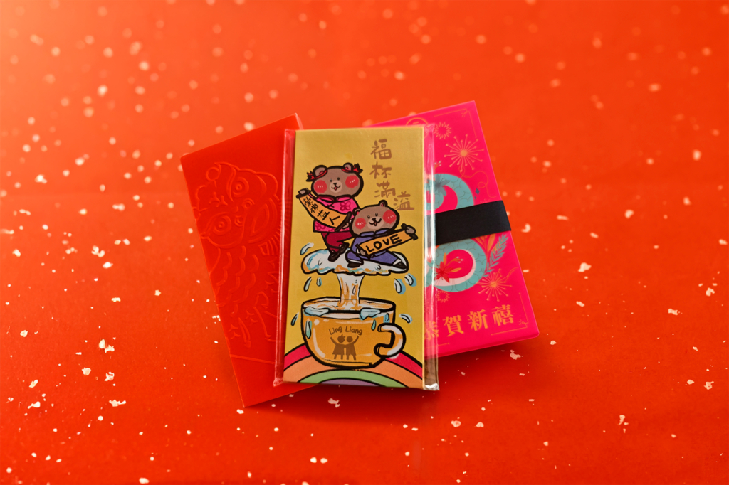 Three lai see (red envelopes) stacked on a wooden table: a yellow cartoon design in a plastic sleeve, a magenta envelope with gold text, and an orange embossed one.
